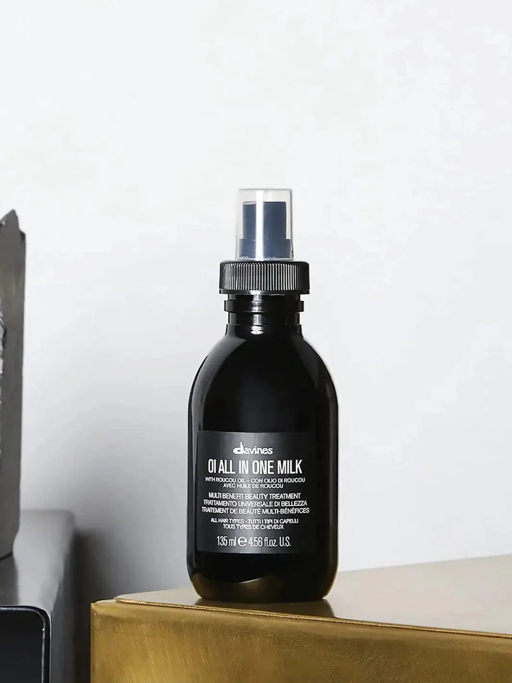 A black spray bottle of Davines OI All in one Milk stands on a metallic gold surface against a plain white wall | Twentyseven Toronto