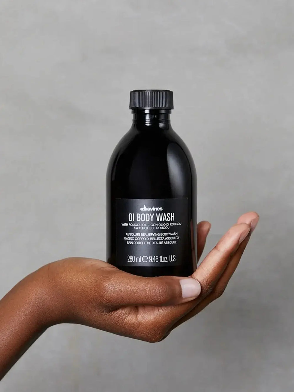 A person holds a glossy black bottle of Davines OI Body Wash upright in the palm of their hand against a textured grey background | Twentyseven Toronto