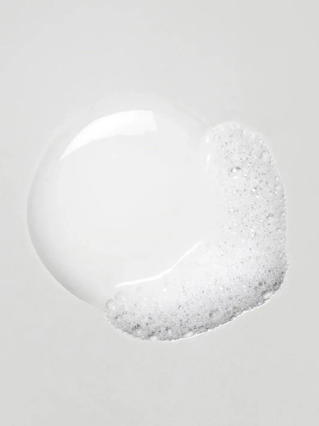A dollop of clear Davines OI Body Wash product forms a smooth puddle with frothy white bubbles on one side against a light grey surface | Twentyseven Toronto