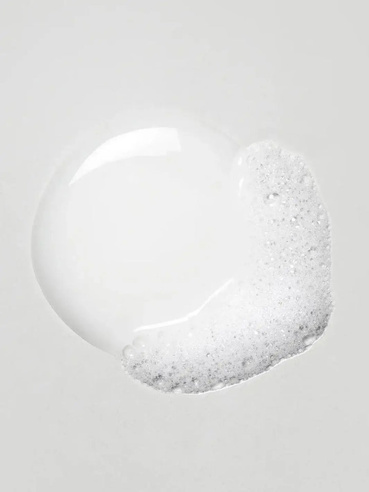 A dollop of clear Davines OI Body Wash product forms a smooth puddle with frothy white bubbles on one side against a light grey surface | Twentyseven Toronto
