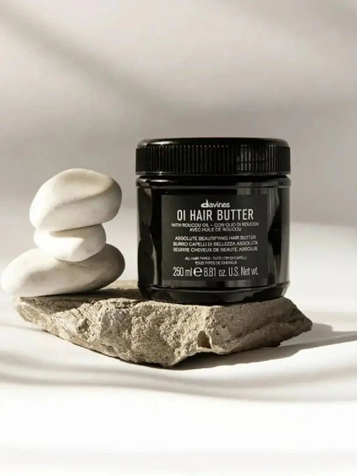 A black jar of Davines OI Hair Butter Mask rests on a grey rock next to a stack of three smooth white balancing stones | Twentyseven Toronto