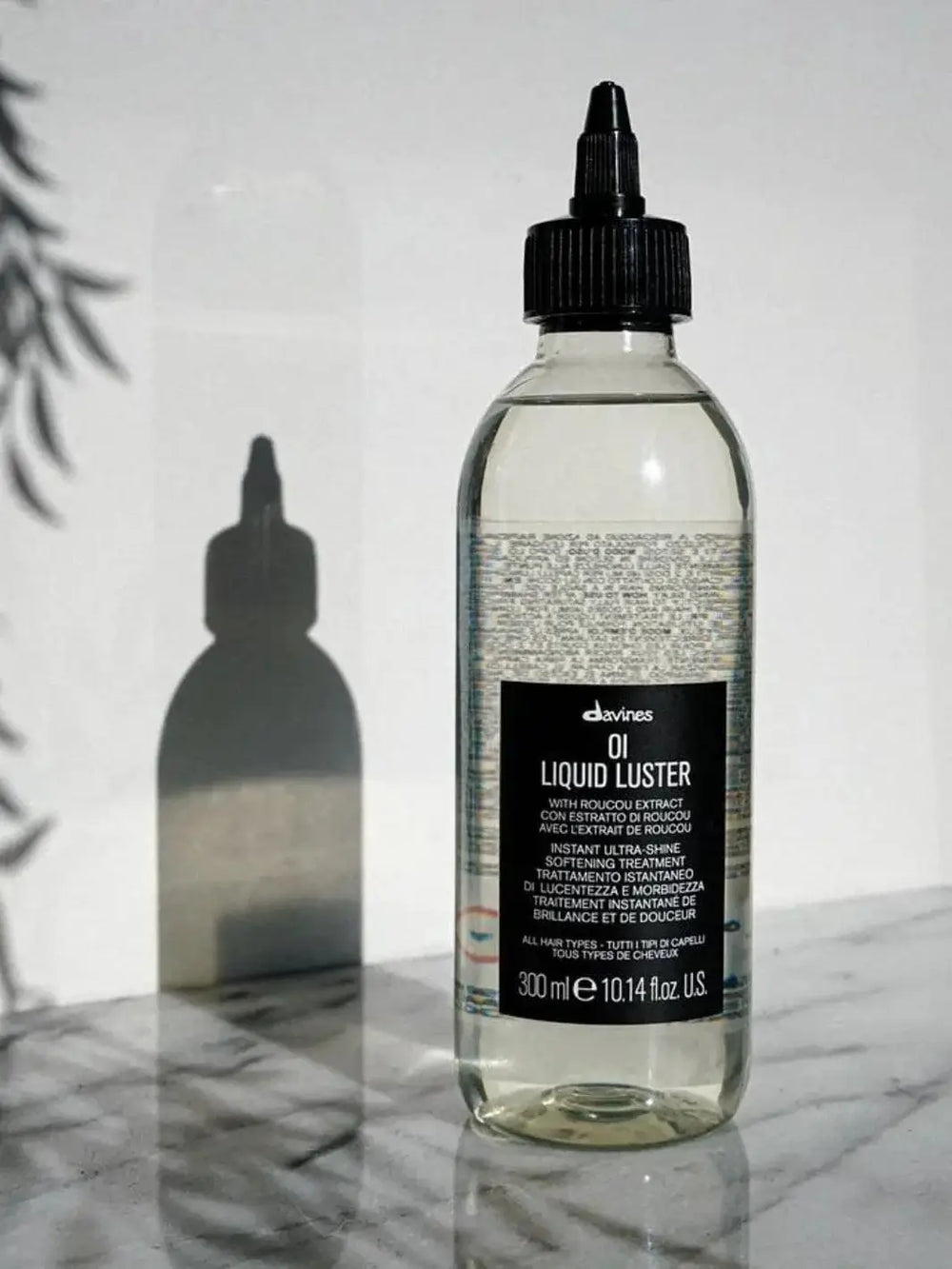 A clear bottle of Davines OI Liquid Lustre stands on a marble surface, casting a shadow against a white wall | Twentyseven Toronto