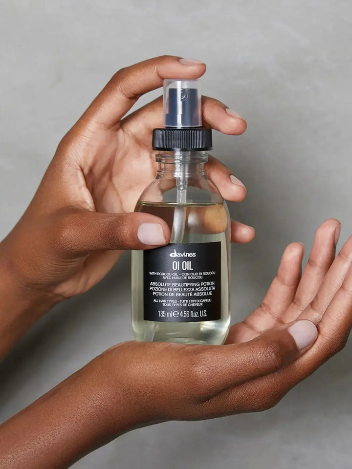 A person holds a clear glass pump bottle of OI Oil Absolute Beautifying Potion upright in their hands against a textured grey background | Twentyseven Toronto