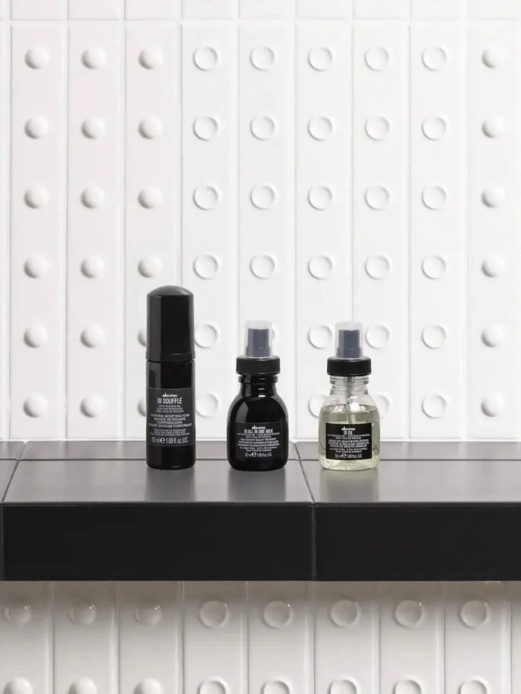 A travel-sized bottle of Davines OI Soufflé stands in a row alongside travel-sized OI All In One Milk and OI Oil on a dark shelf against a textured white wall | Twentyseven Toronto