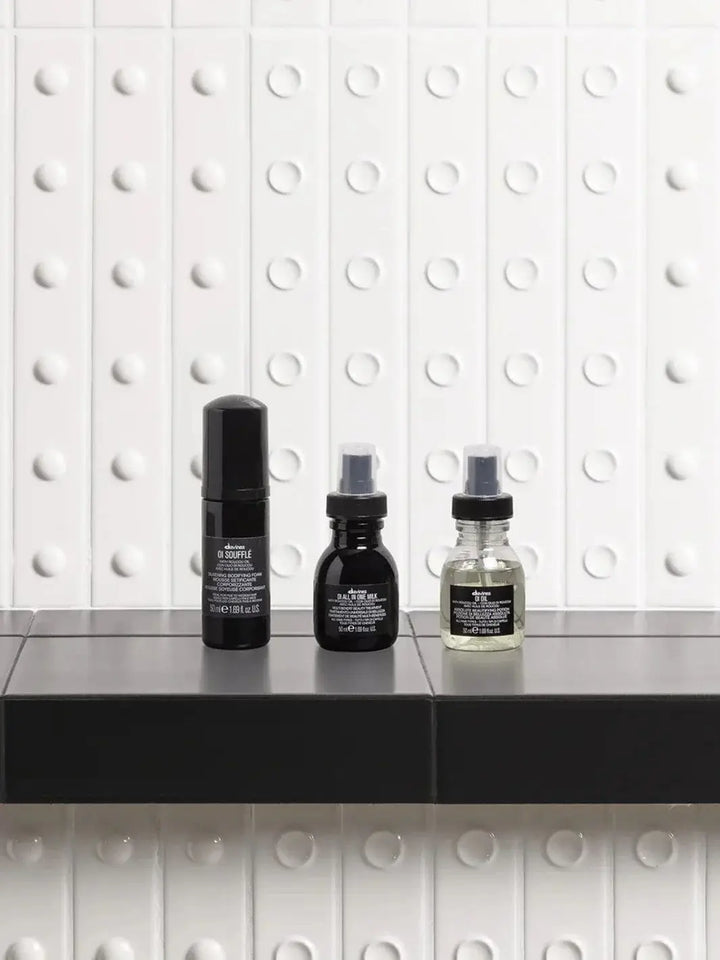 A travel-sized bottle of Davines OI Soufflé stands in a row alongside travel-sized OI All In One Milk and OI Oil on a dark shelf against a textured white wall | Twentyseven Toronto