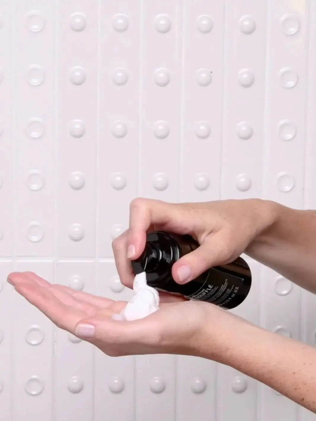 A person's hands dispense a white foam from a bottle of Davines OI Soufflé against a white tiled wall with circular accents | Twentyseven Toronto