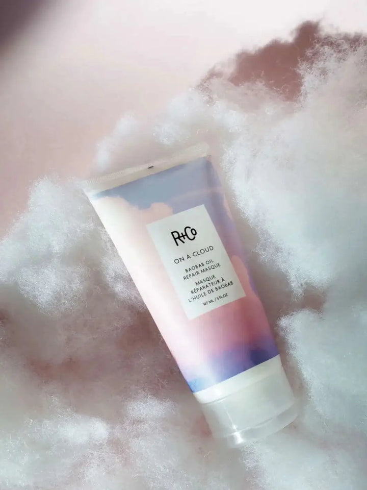 A pastel-colored tube of R+Co ON A CLOUD Repair Mask rests gently on a bed of fluffy, white cloud-like material against a soft pink background | Twentyseven Toronto
