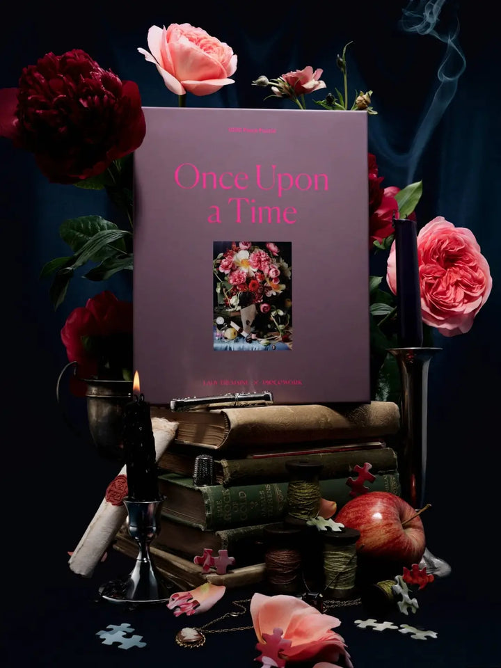 Purple jigsaw puzzle packaging titled 'Once Upon a Time' 1000 piece puzzle by Piecework Puzzles on top of a stack of books and surrounded by vintage decor and flowers | Twentyseven Toronto