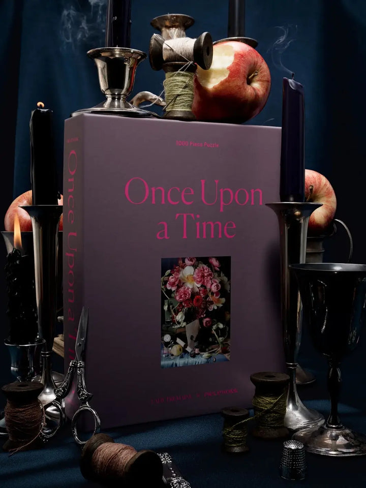 Purple jigsaw puzzle packaging titled 'Once Upon a Time' 1000 piece puzzle from Piecework Puzzles surrounded by vintage props on a dark background | Twentyseven Toronto