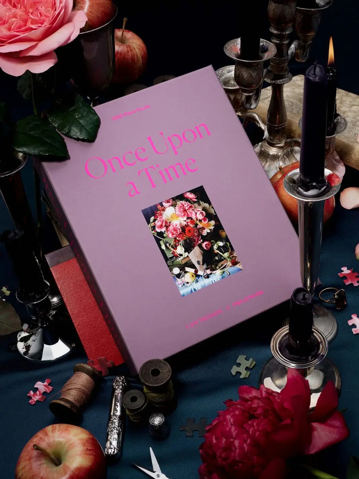 Purple jigsaw puzzle packaging titled 'Once Upon a Time' 1000 piece puzzle from Piecework Puzzles surrounded by candles and flowers on a dark surface | Twentyseven Toronto