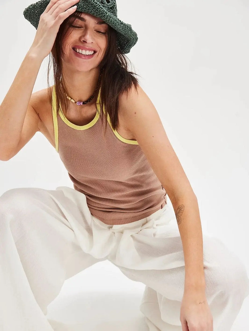 A model smiles while crouching in the Free People Only 1 Solid Ringer Tank - Praline Sunshine Combo, styled with loose white pants and a green crochet bucket hat | Twentyseven Toronto