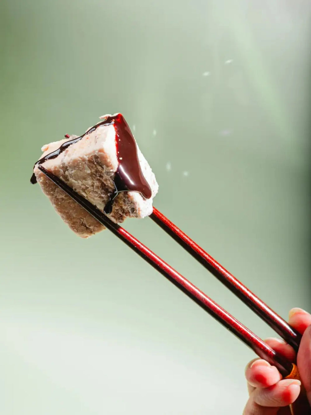 A pair of dark wooden chopsticks to lift a single, square-shaped Mojave Mallows Organic S'more Speckled Marshmallow drizzled with thick chocolate syrup against a soft green background | Twentyseven Toronto