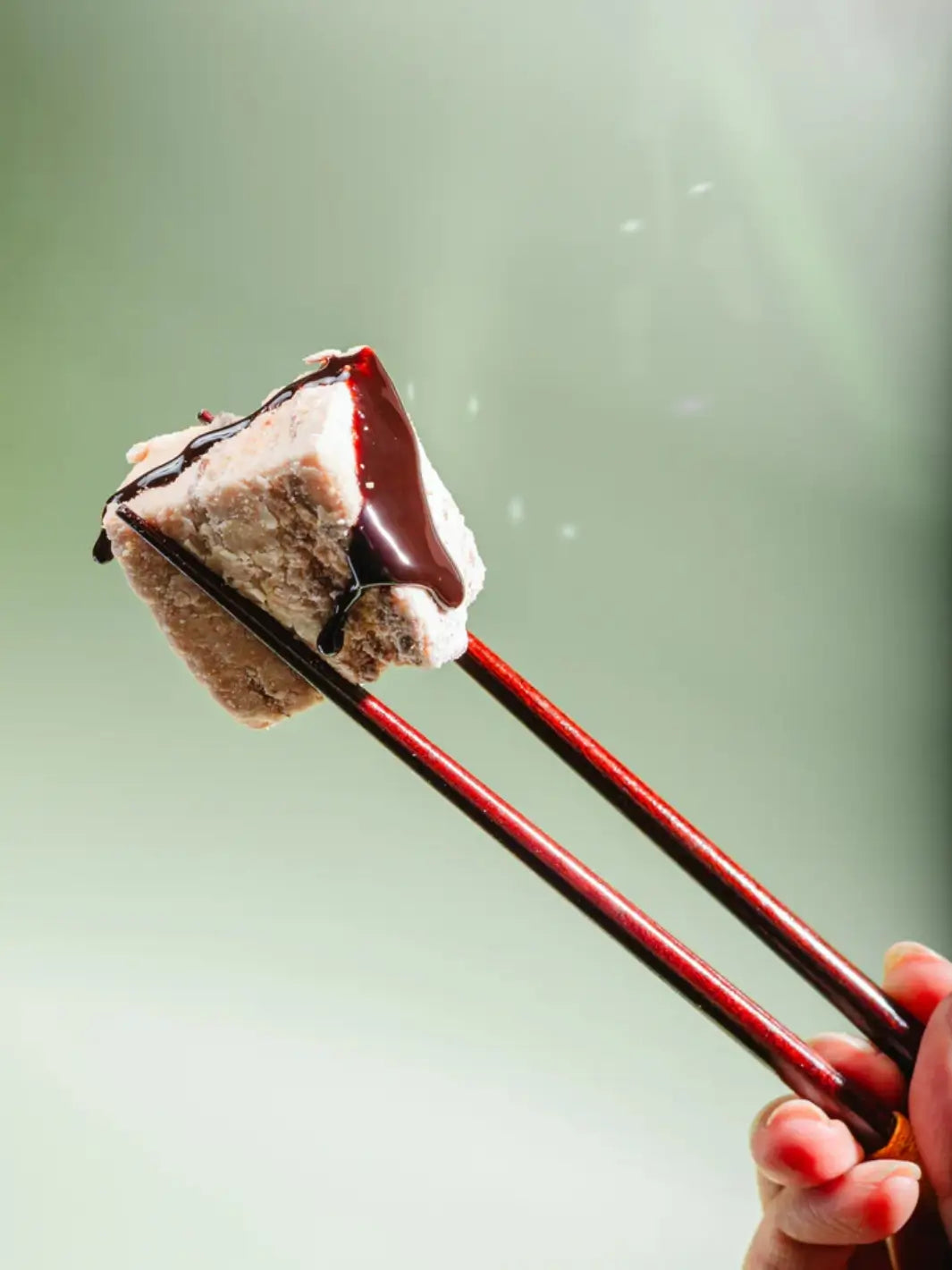 A pair of dark wooden chopsticks to lift a single, square-shaped Mojave Mallows Organic S'more Speckled Marshmallow drizzled with thick chocolate syrup against a soft green background | Twentyseven Toronto