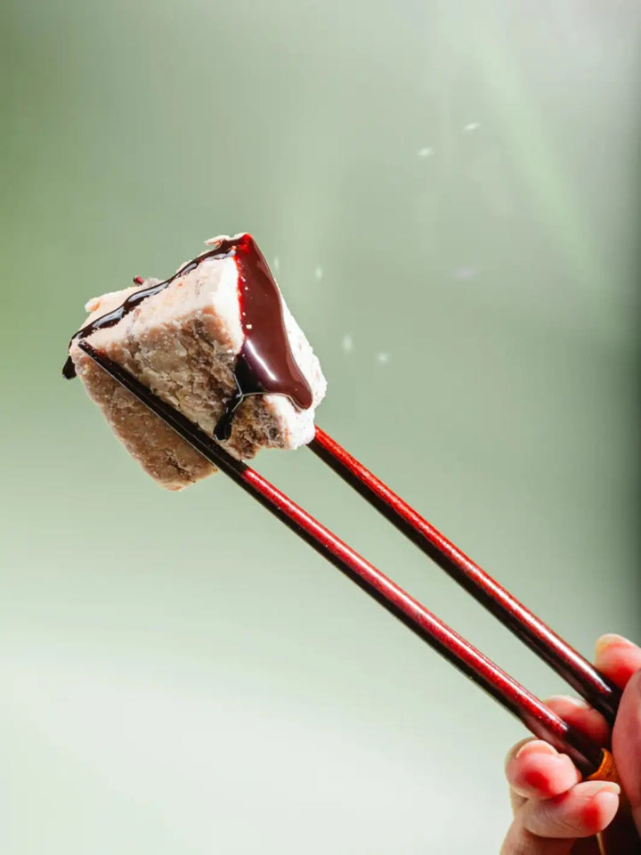 A pair of dark wooden chopsticks to lift a single, square-shaped Mojave Mallows Organic S'more Speckled Marshmallow drizzled with thick chocolate syrup against a soft green background | Twentyseven Toronto