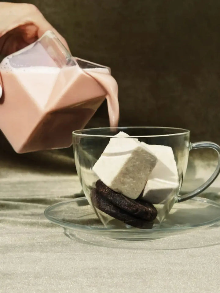 A hand pours chocolate milk from a geometric glass carton into a clear mug filled with cookies and Mojave Mallows Organic Vanilla Bean Marshmallows | Twentyseven Toronto