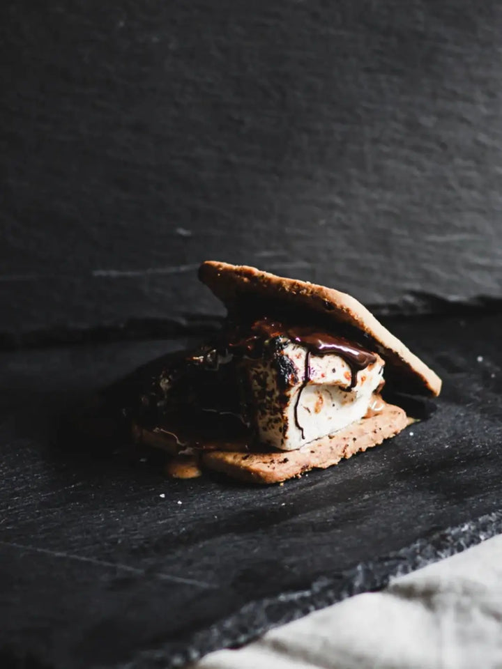 A fully assembled s'more featuring a toasted marshmallow, melted chocolate, and graham crackers against a dark slate background, highlighting Mojave Mallows Organic Vanilla Bean Marshmallows | Twentyseven Toronto