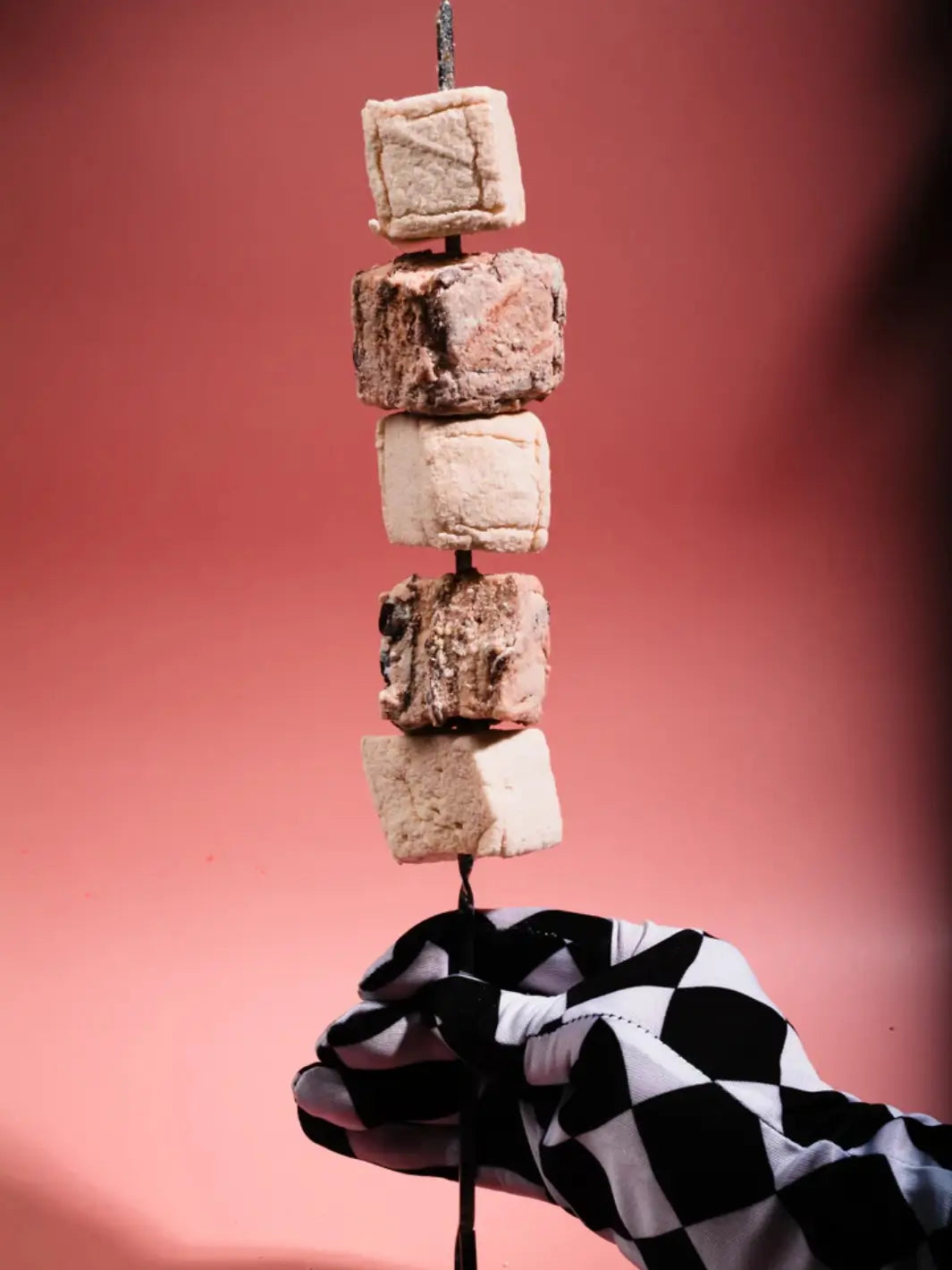 A hand wearing a black-and-white checkered glove holds a skewer stacked with Mojave Mallows Organic Vanilla Bean Marshmallows against a pink background | Twentyseven Toronto