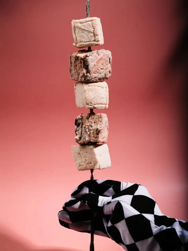 A hand wearing a black-and-white checkered glove holds a skewer stacked with Mojave Mallows Organic Vanilla Bean Marshmallows against a pink background | Twentyseven Toronto