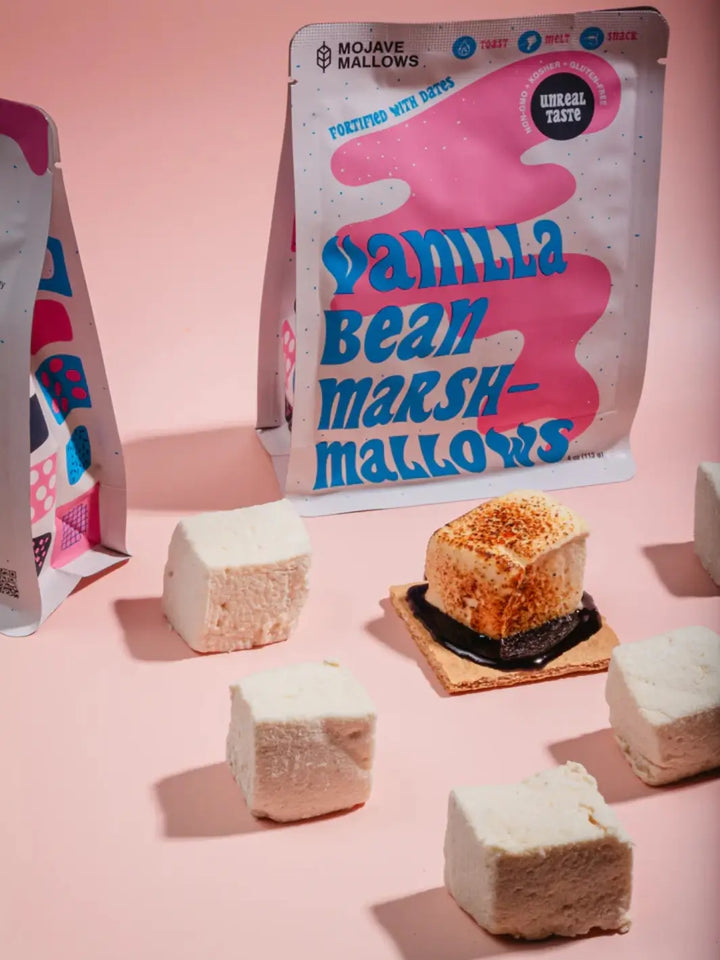 Several square-cut Mojave Mallows Organic Vanilla Bean Marshmallows and a small prepared s'more sit on a pink surface in front of their upright product packaging | Twentyseven Toronto