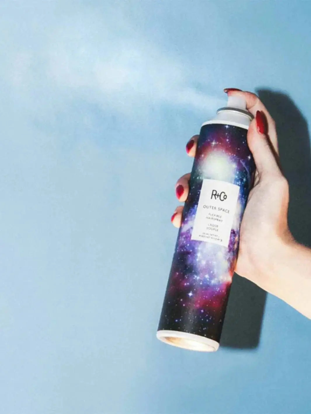 A hand with red nails sprays a fine mist from a can of R+Co OUTER SPACE Flexible Hairspray against a solid blue background | Twentyseven Toronto