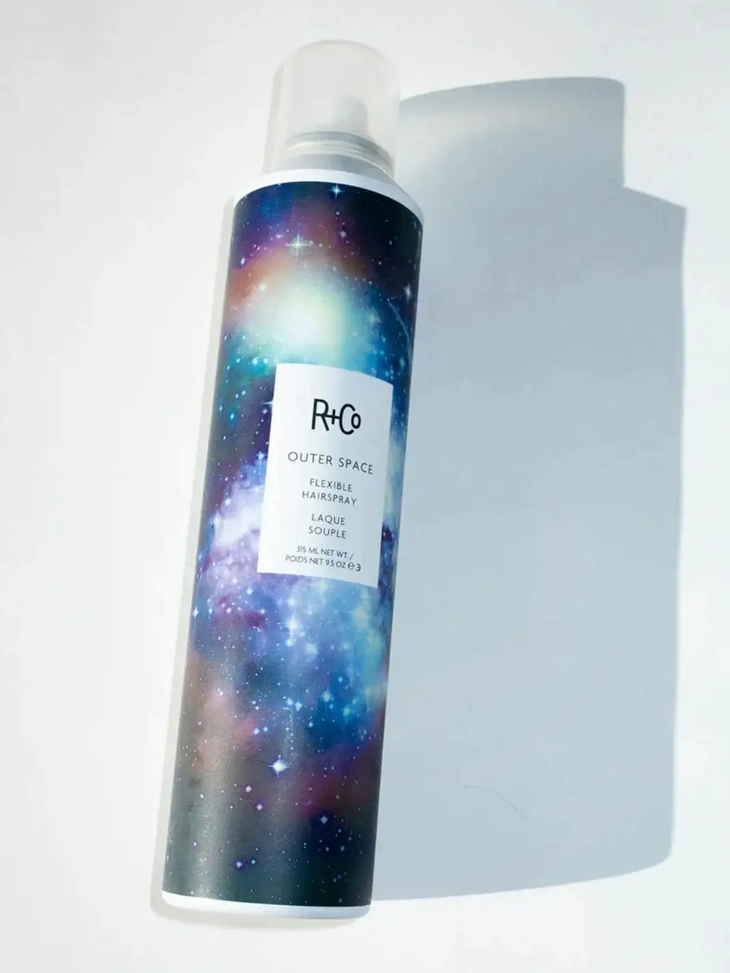 A full-size aluminum can of R+Co OUTER SPACE Flexible Hairspray with a colorful galaxy print label rests on a bright white surface | Twentyseven Toronto