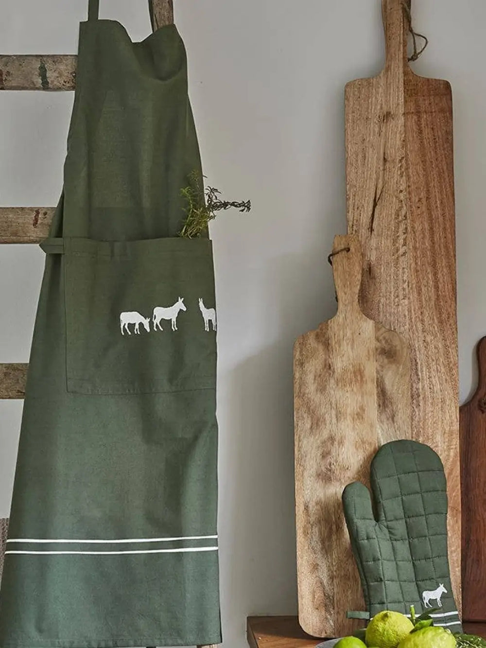 Green apron with farm animals design hanging on a wooden rack with cutting boards and oven mitts from Sylvie Thiriez | Twentyseven Toronto
