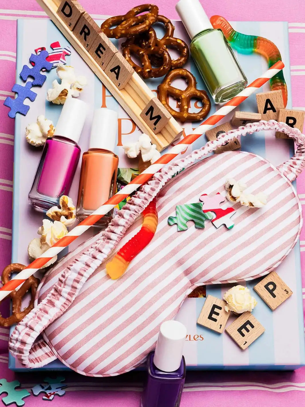 Colorful items including nail polish, a sleep mask, and snacks on a pink background atop Piecework Puzzles 'Pajama Party' 1000 piece puzzle | Twentyseven Toronto