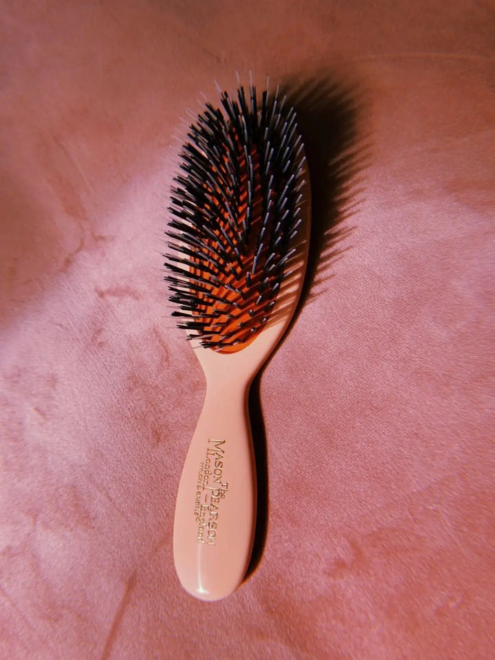 A premium pink hairbrush with mixed boar and nylon bristles on a red rubber cushion is showcased against a soft pink velvet background, emphasizing the luxury design of the Mason Pearson Pastel Pink Boar Bristle & Nylon Mix Hair Brush | Twentyseven Toronto