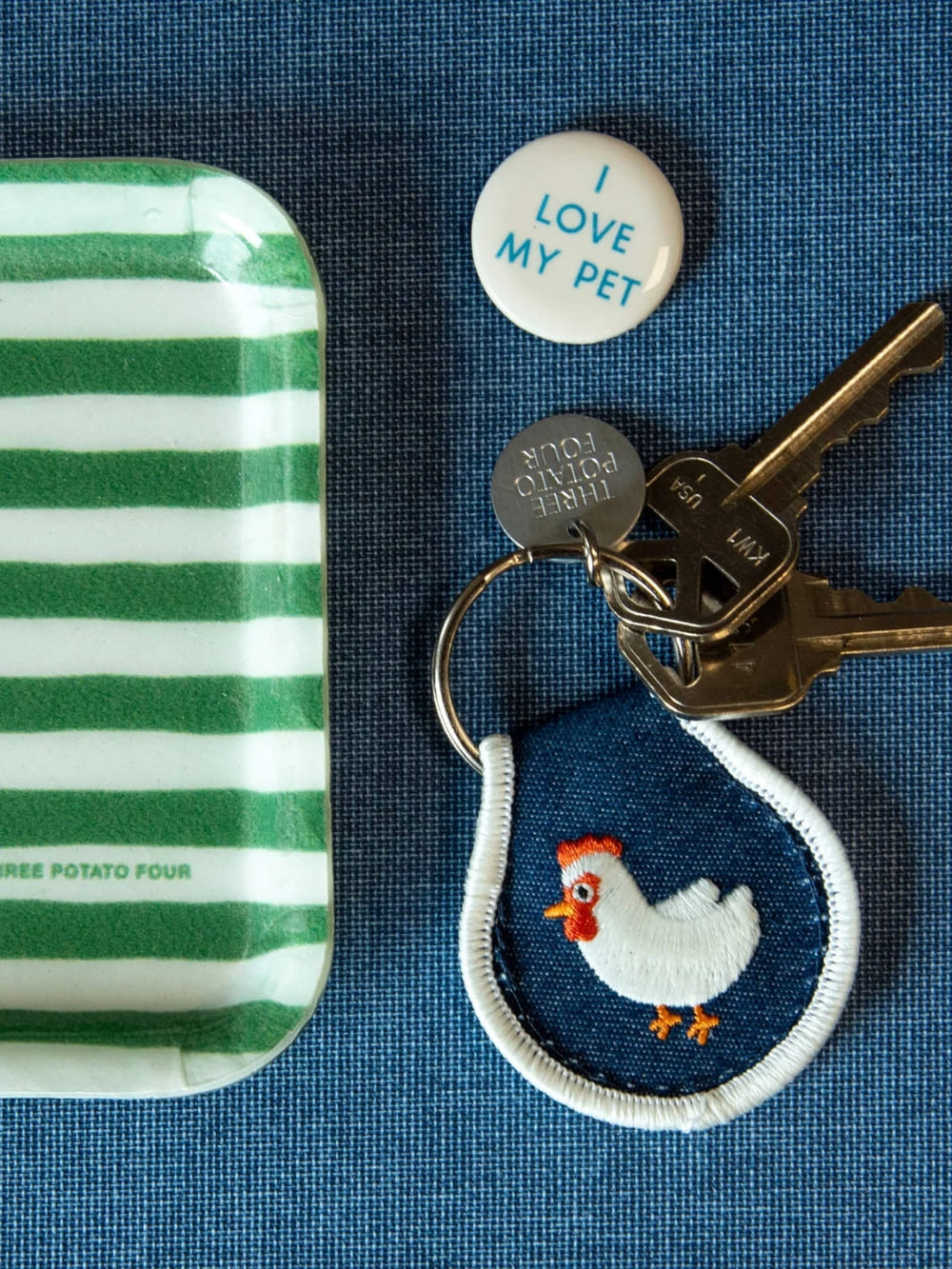 Patch keychain with embroidered chicken design from Three Potato Four next to green and white striped tray, and 'I Love My Pet' button on blue background | Twentyseven Toronto