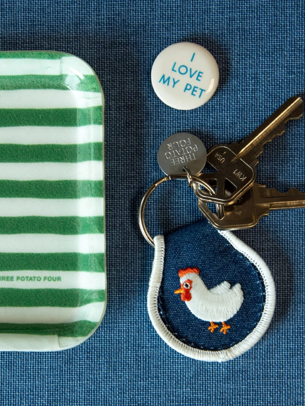 Patch keychain with embroidered chicken design from Three Potato Four next to green and white striped tray, and 'I Love My Pet' button on blue background | Twentyseven Toronto