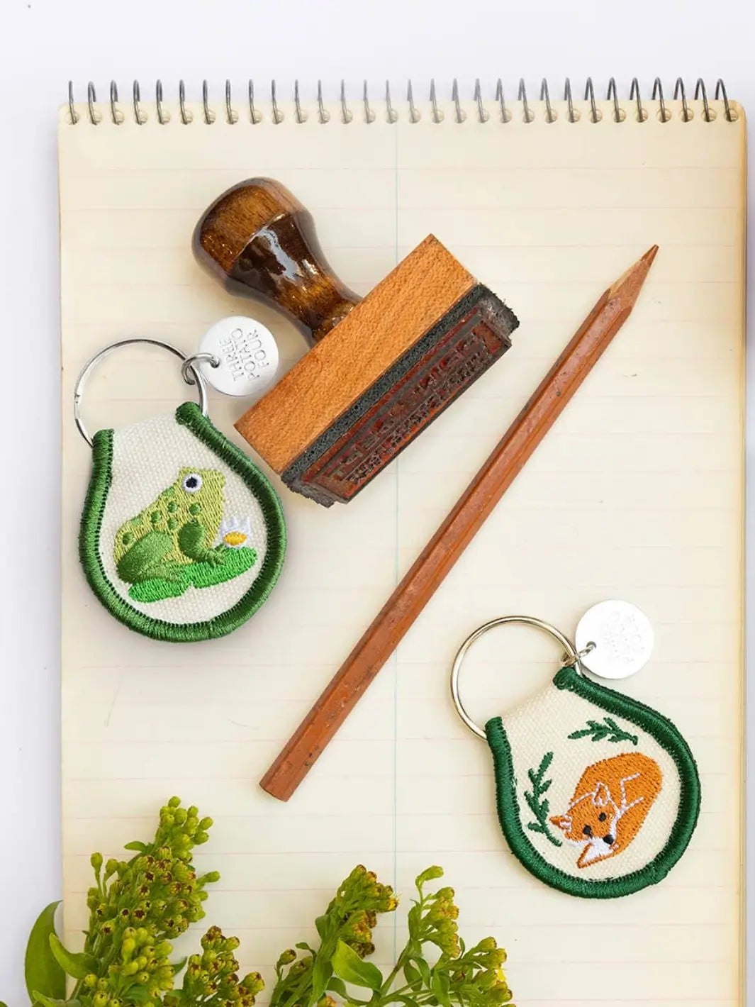 A Three Potato Four Patch Keychain - Frog rests on a yellowed notebook alongside a wooden stamp, a pencil, and a matching fox-themed patch keychain | Twentyseven Toronto