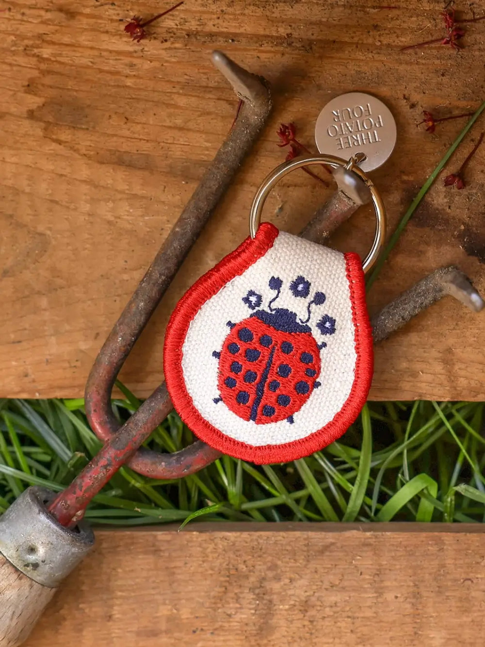 A Three Potato Four Patch Keychain - Ladybug rests on a rustic metal garden hand rake over green grass and a wooden box | Twentyseven Toronto