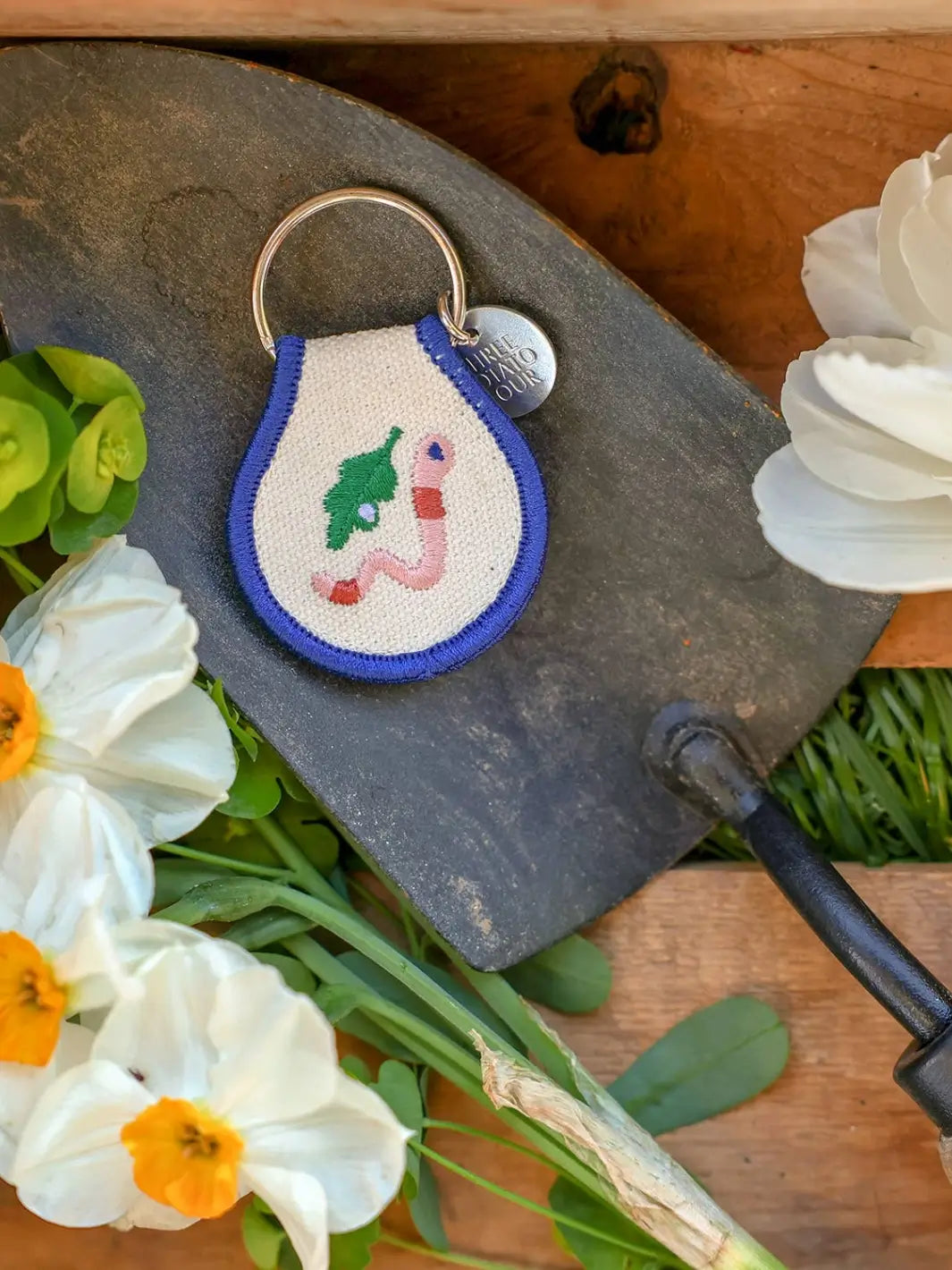 A Three Potato Four Patch Keychain - Worm sits on a dark metal garden trowel surrounded by fresh white and yellow flowers | Twentyseven Toronto
