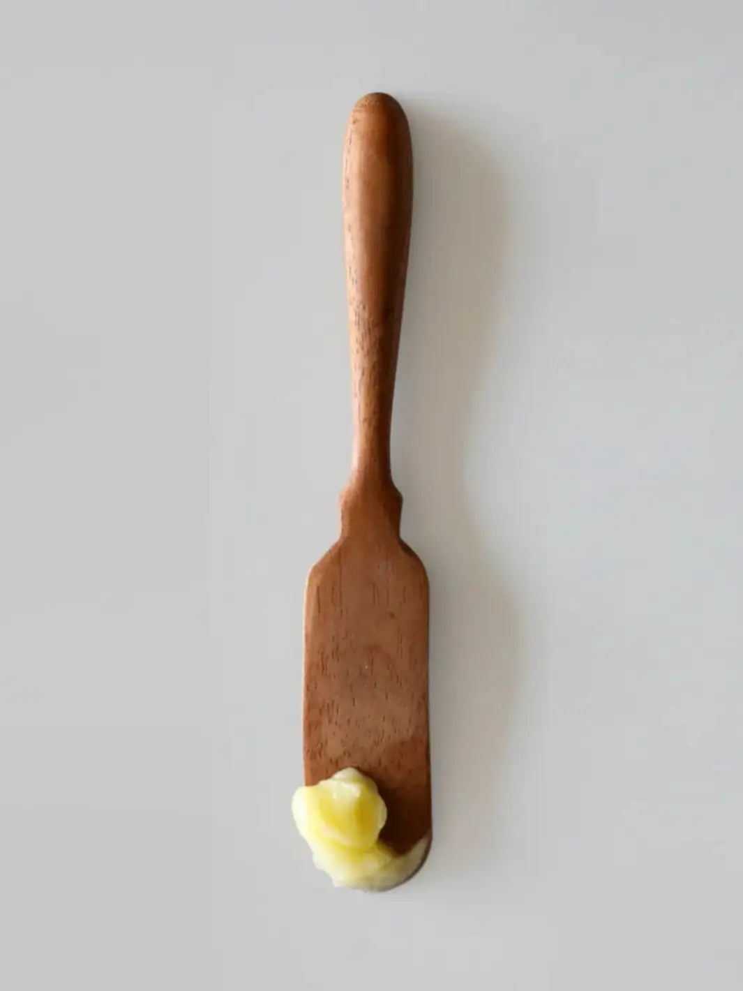 A small dollop of yellowish Jao Brand Patio Oil product sits on the tip of a vertical wooden spatula over a plain white background | Twentyseven Toronto