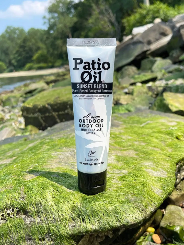 A squeeze tube of Jao Brand Patio Oil Sunset Blend stands upright on a bright green, moss-covered rock near a body of water | Twentyseven Toronto