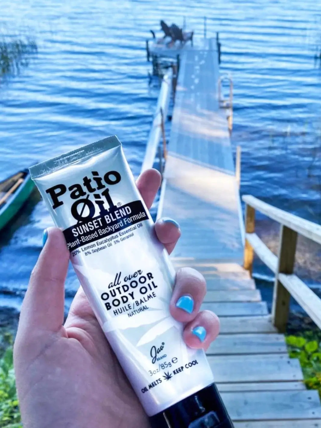 A hand with blue nail polish holds up a tube of Jao Brand Patio Oil Sunset Blend with a long wooden dock and calm blue lake water in the background | Twentyseven Toronto