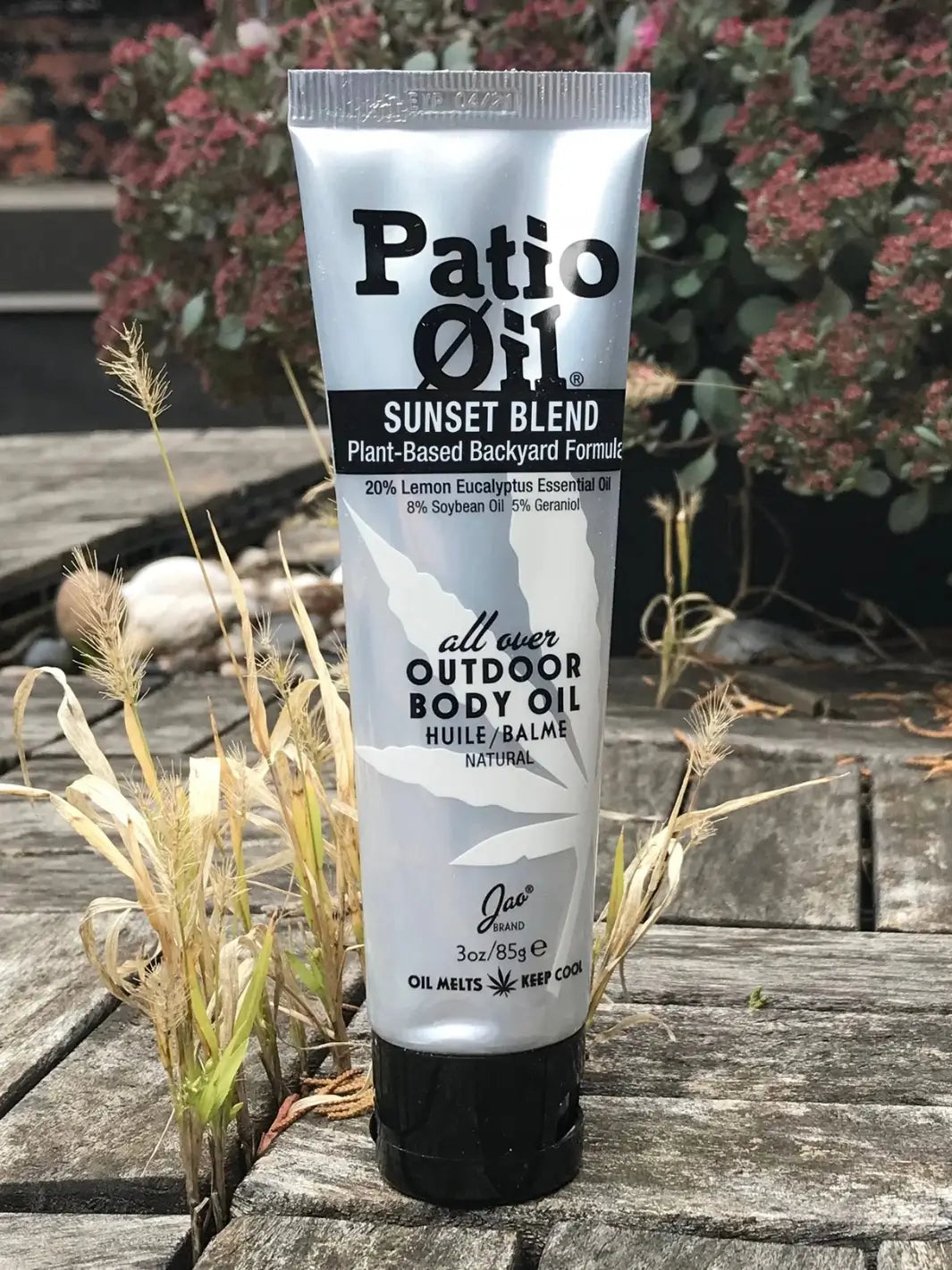 A squeeze tube of Jao Brand Patio Oil Sunset Blend sits on a weathered wooden deck surrounded by small tufts of dry grass | Twentyseven Toronto