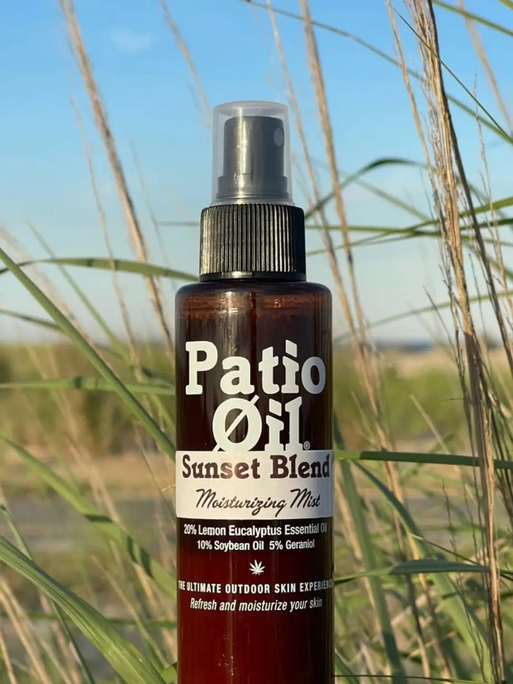 A close-up shot of a bottle of Jao Brand Patio Oil Moisturizing Mist set outdoors against a background of tall green grass and a blue sky | Twentyseven Toronto