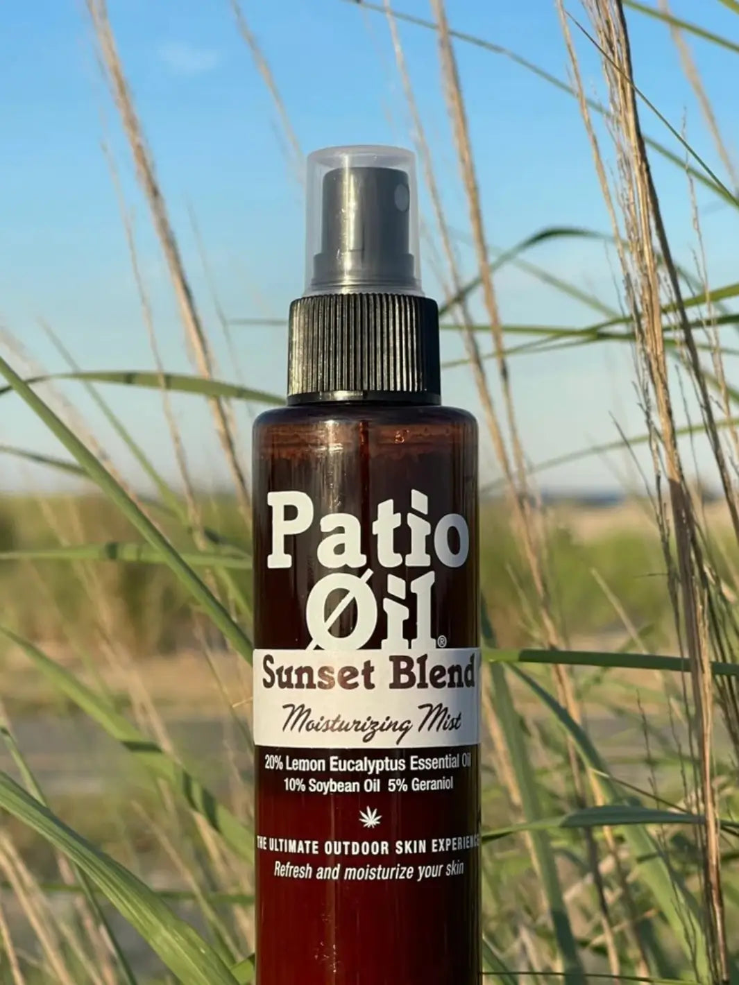 A close-up shot of a bottle of Jao Brand Patio Oil Moisturizing Mist set outdoors against a background of tall green grass and a blue sky | Twentyseven Toronto