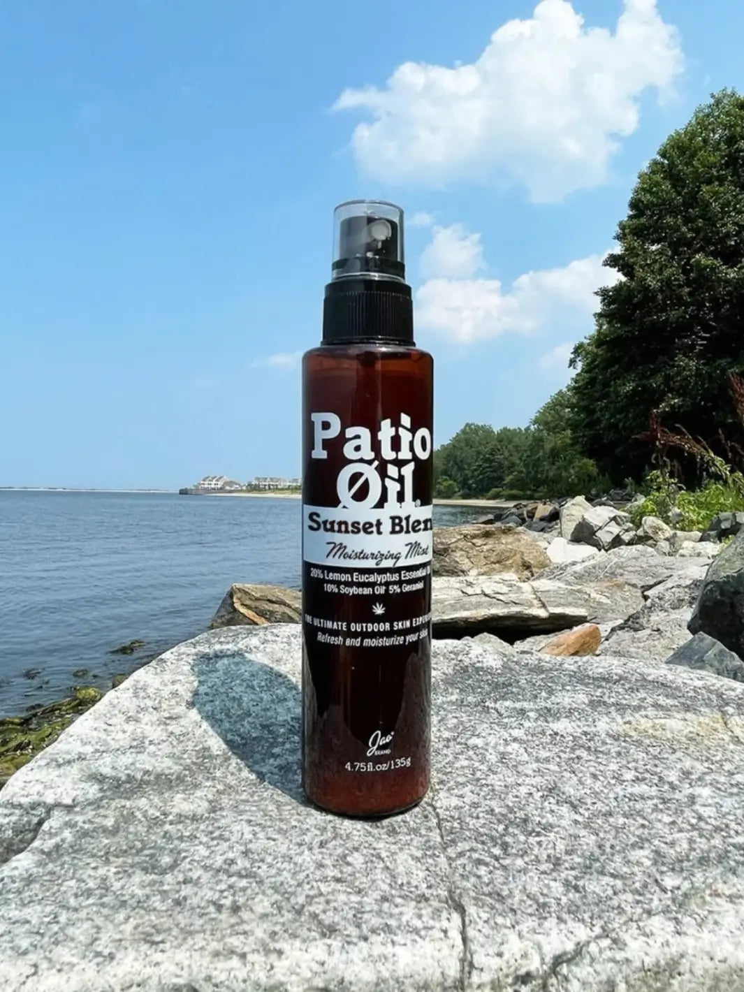 A bottle of Jao Brand Patio Oil Moisturizing Mist stands upright on a large gray boulder along a rocky shoreline next to the ocean | Twentyseven Toronto