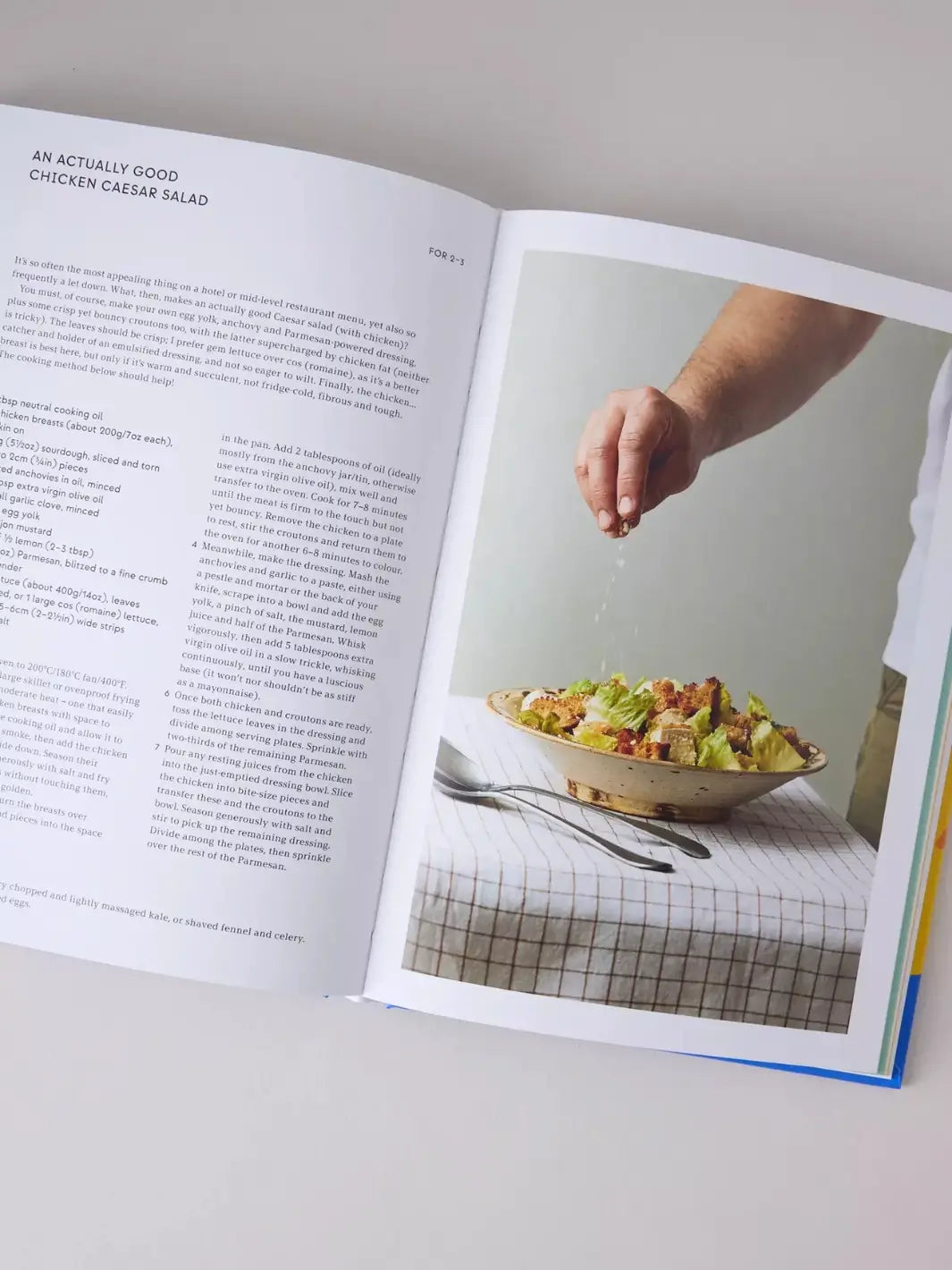 Page from the cookbook Peckish: An Inspirational Collection of Winning Chicken Dinners by Ed Smith features a recipe titled "An Actually Good Chicken Caesar Salad" alongside a full-page photo of a hand seasoning a fresh salad in a ceramic bowl | Twentyseven Toronto