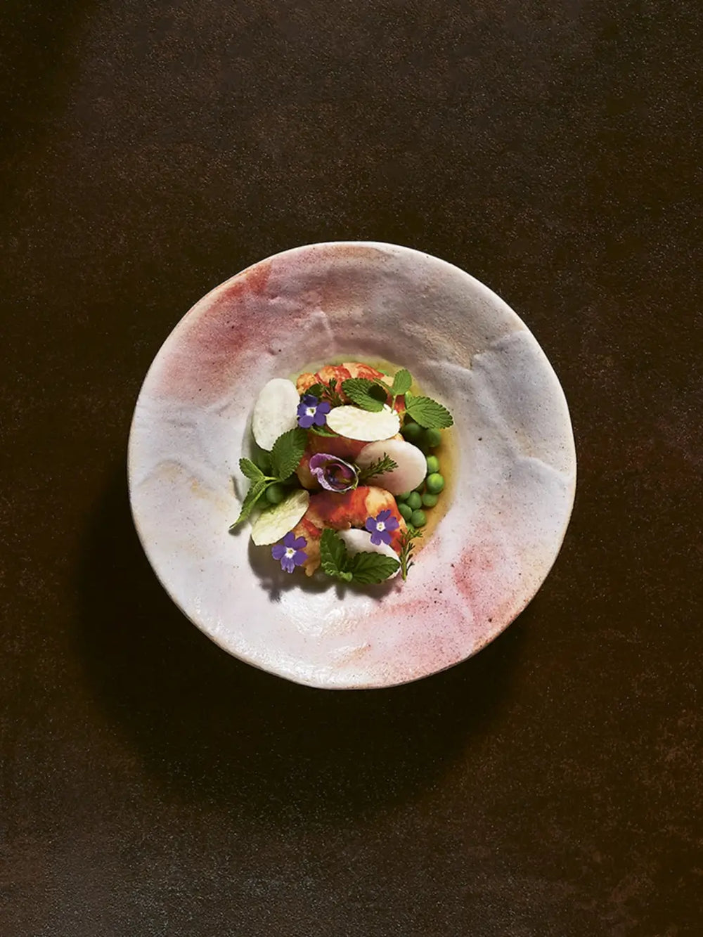 A dish from Perch: Soil / Land / Sea - A Cookbook by Justin Champagne-Lagarde & Mandel Hitzer features a vibrant seafood dish served in a textured, pink-hued ceramic bowl, garnished with fresh green peas, mint leaves, white radish slices, and purple edible flowers | Twentyseven Toronto
