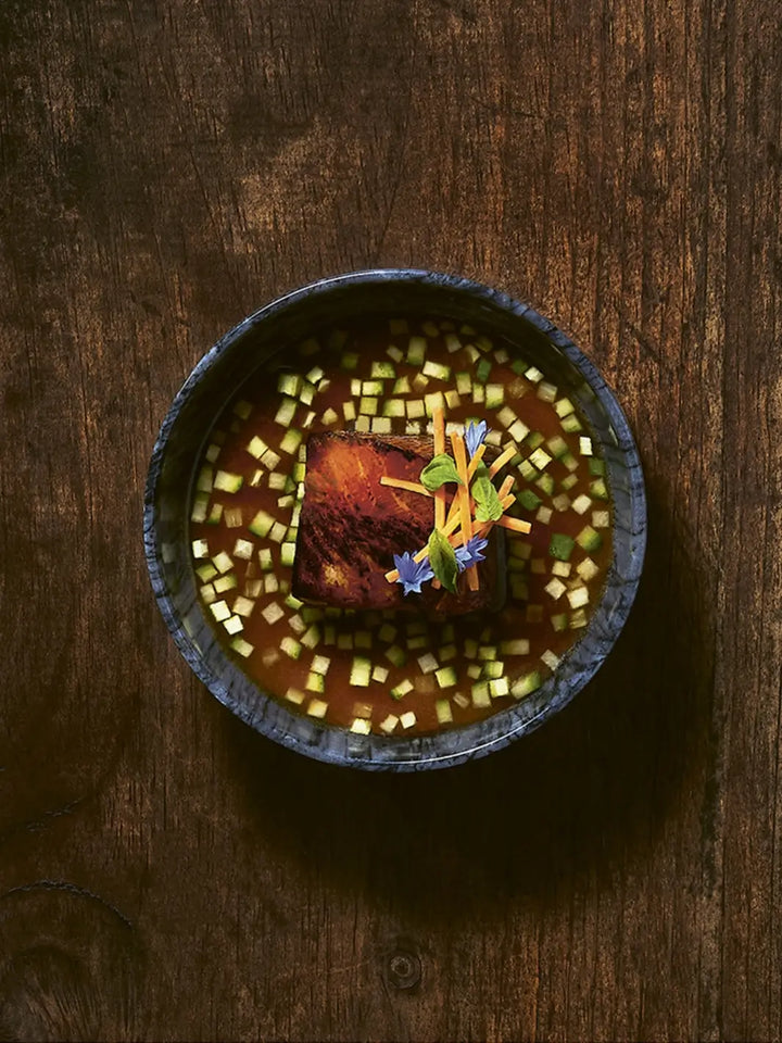A dish from Perch: Soil / Land / Sea - A Cookbook by Justin Champagne-Lagarde & Mandel Hitzer showcasing a dark, seared protein centered in a blue marble-patterned bowl, surrounded by broth filled with finely diced green vegetables | Twentyseven Toronto