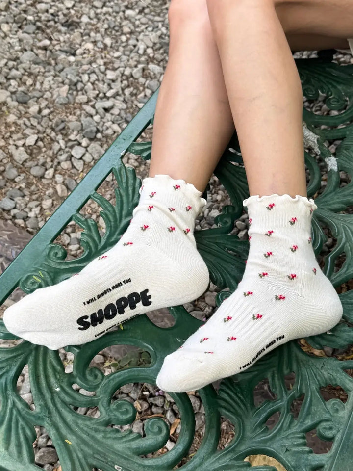A side view shows a person sitting on a wrought-iron bench wearing the Le Bon Shoppe Petal Ruffled Hugger Crew Socks in Cream Petal | Twentyseven