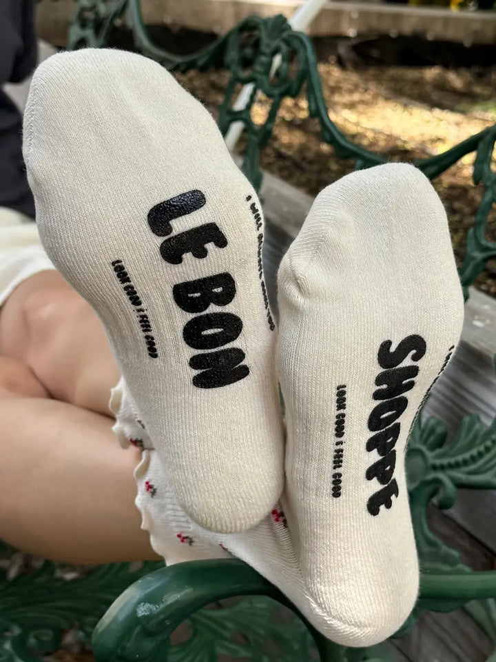 A close-up view shows the bottoms of the Le Bon Shoppe Petal Ruffled Hugger Crew Socks in Cream Petal with black brand lettering on the soles | Twentyseven