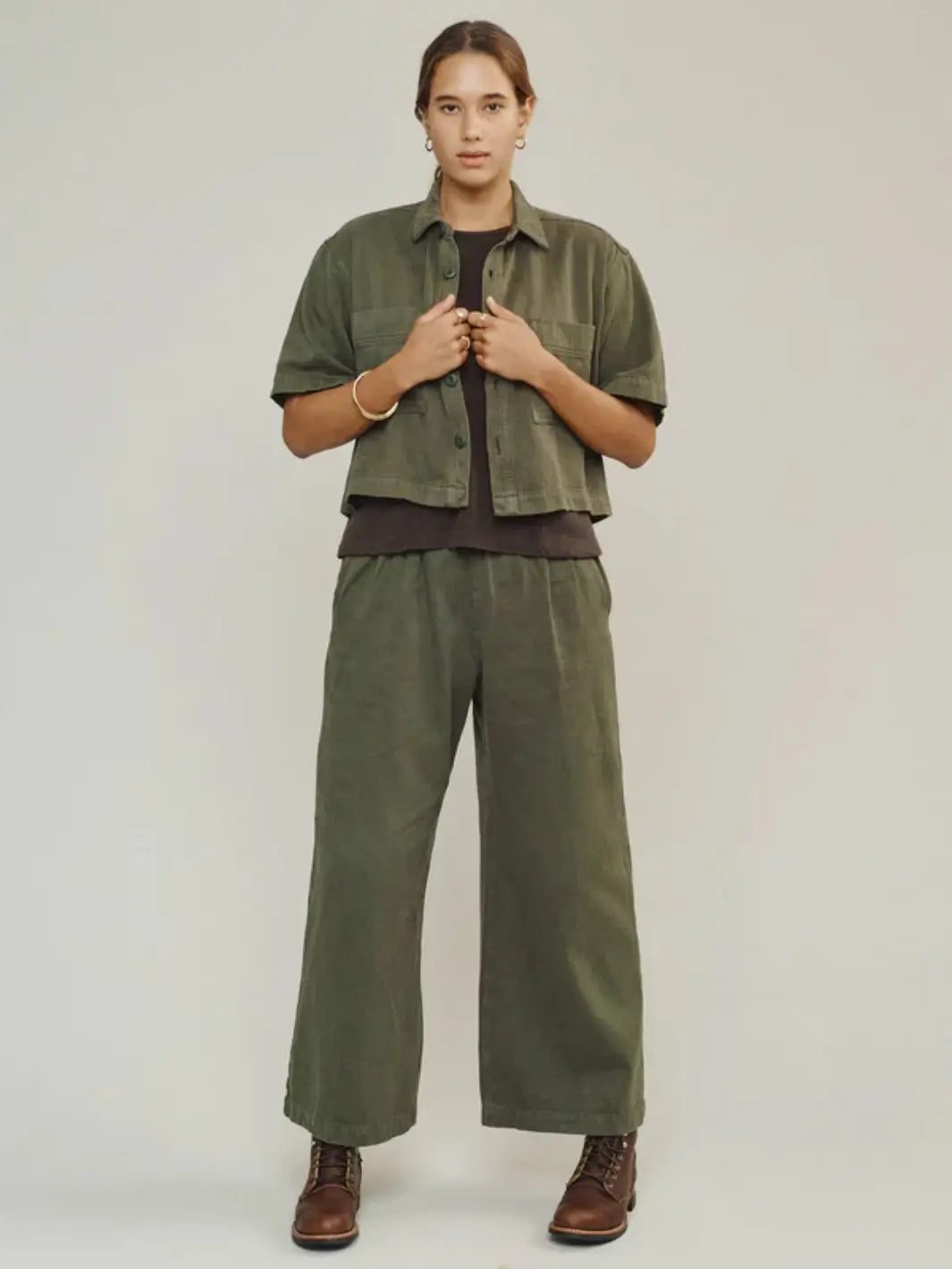 A full-length front view shows a model wearing the Jungmaven Phinney Twill Shirt - Olive Green open over a dark t-shirt with matching pants and boots | Twentyseven Toronto