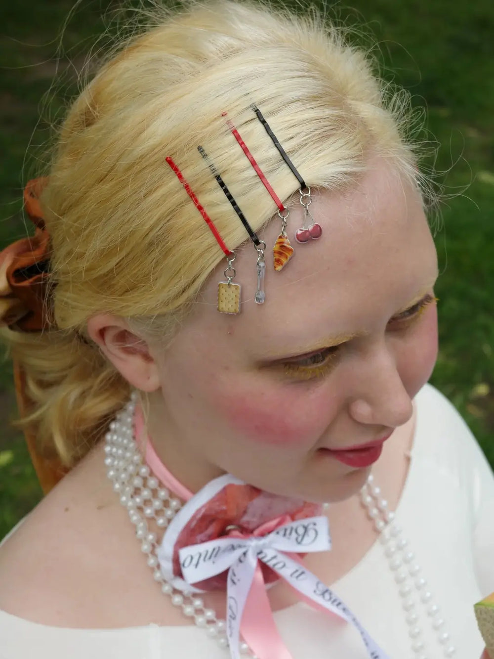 A person with blonde hair wears four alternating red and black Chunks Hairclips Picnic Bobby Pins, each featuring a dangling miniature charm of a cracker, a fork, a croissant, and cherries | Twentyseven Toronto