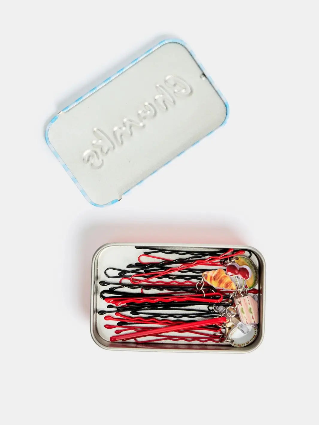 An open, rectangular metal tin shows a collection of black and red Chunks Hairclips Picnic Bobby Pins stored inside alongside the reverse side of the lid featuring an embossed brand logo | Twentyseven Toronto