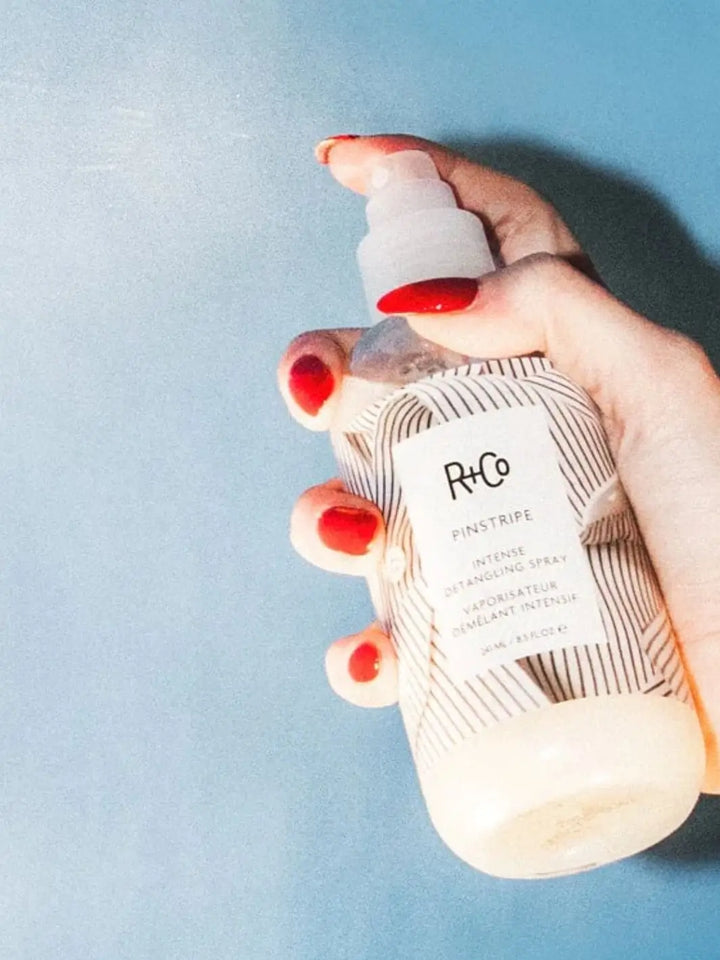 A hand with bright red fingernails presses the nozzle of a bottle to spray R+Co PINSTRIPE Intense Detangling Spray against a light blue background | Twentyseven Toronto