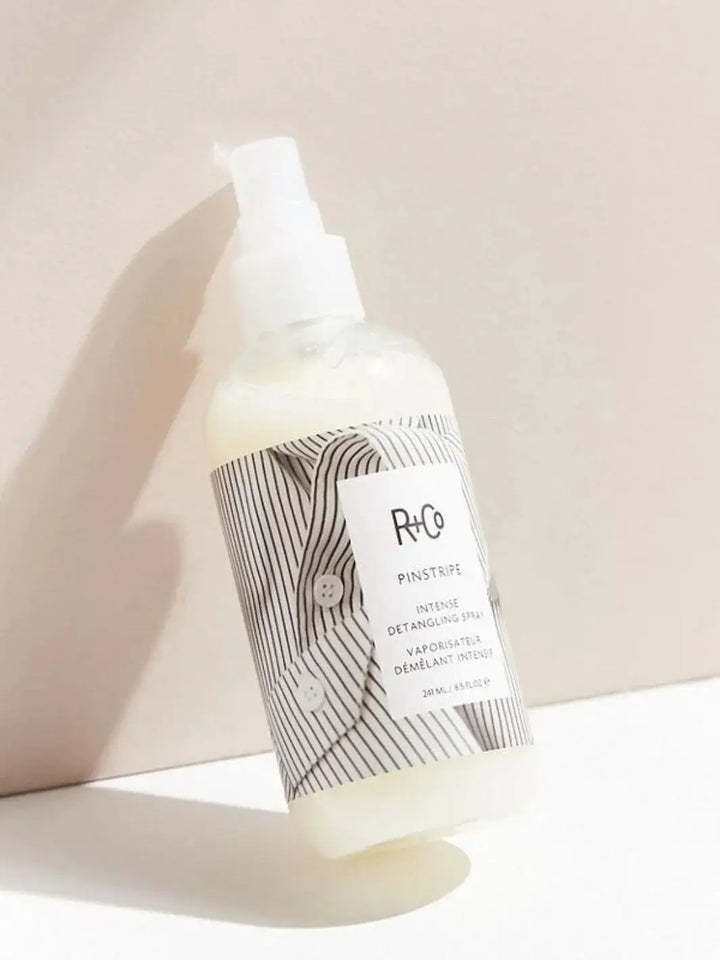 A bottle of R+Co PINSTRIPE Intense Detangling Spray features a striped shirt design on its label while resting against a neutral, warm background | Twentyseven Toronto